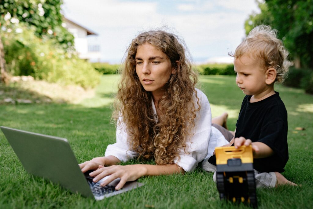 Woman working on a laptop while lying on grass with her child nearby playing. Earn Extra Cash