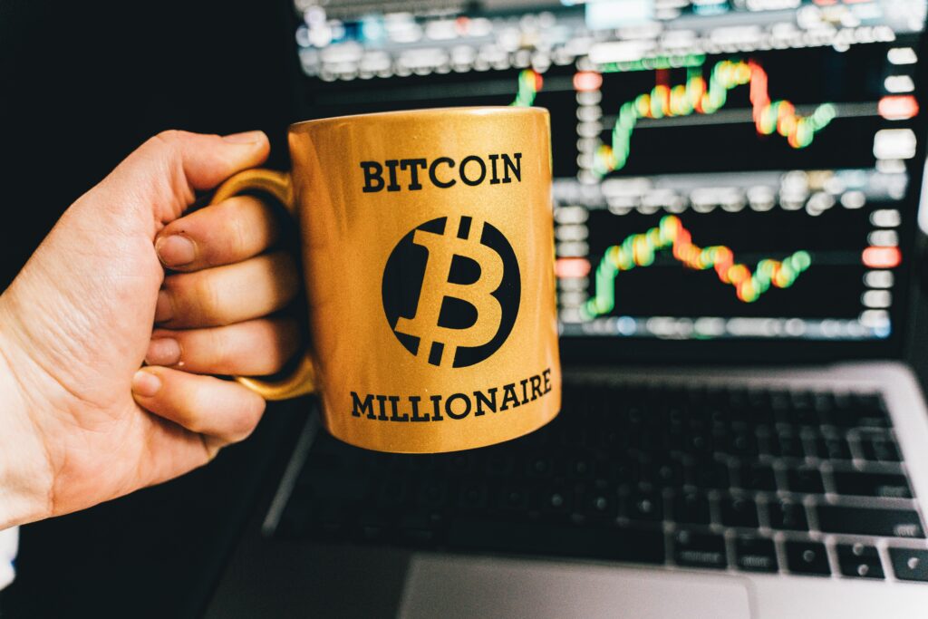 Golden mug with 'Bitcoin Millionaire' text in front of stock market charts on laptop screen. Investing in Malaysia Made Simple: 5 Myths Debunked