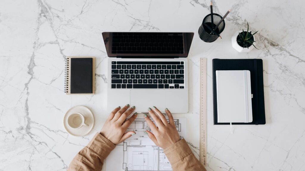 Top view of a stylish home office desk with a laptop, planner, and coffee cup, showing hands on a blueprint. The Truth About Freelancing