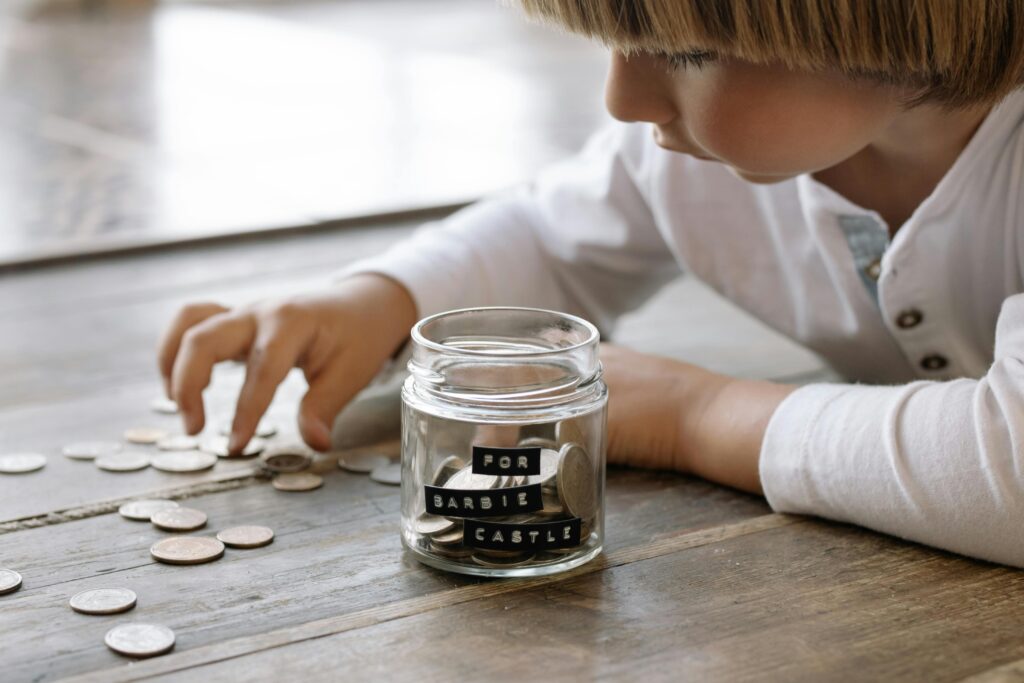 A young child collects coins in a jar labeled 'For Barbie Castle', symbolizing saving and dreams. Teach Kids About Money