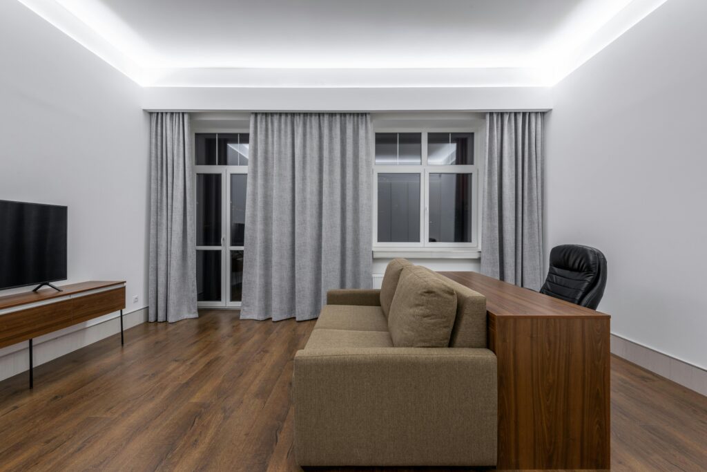 Wooden table near comfortable sofa against modern TV in spacious living room with curtains on window and minimalist design at home. Minimalist Living in Malaysia