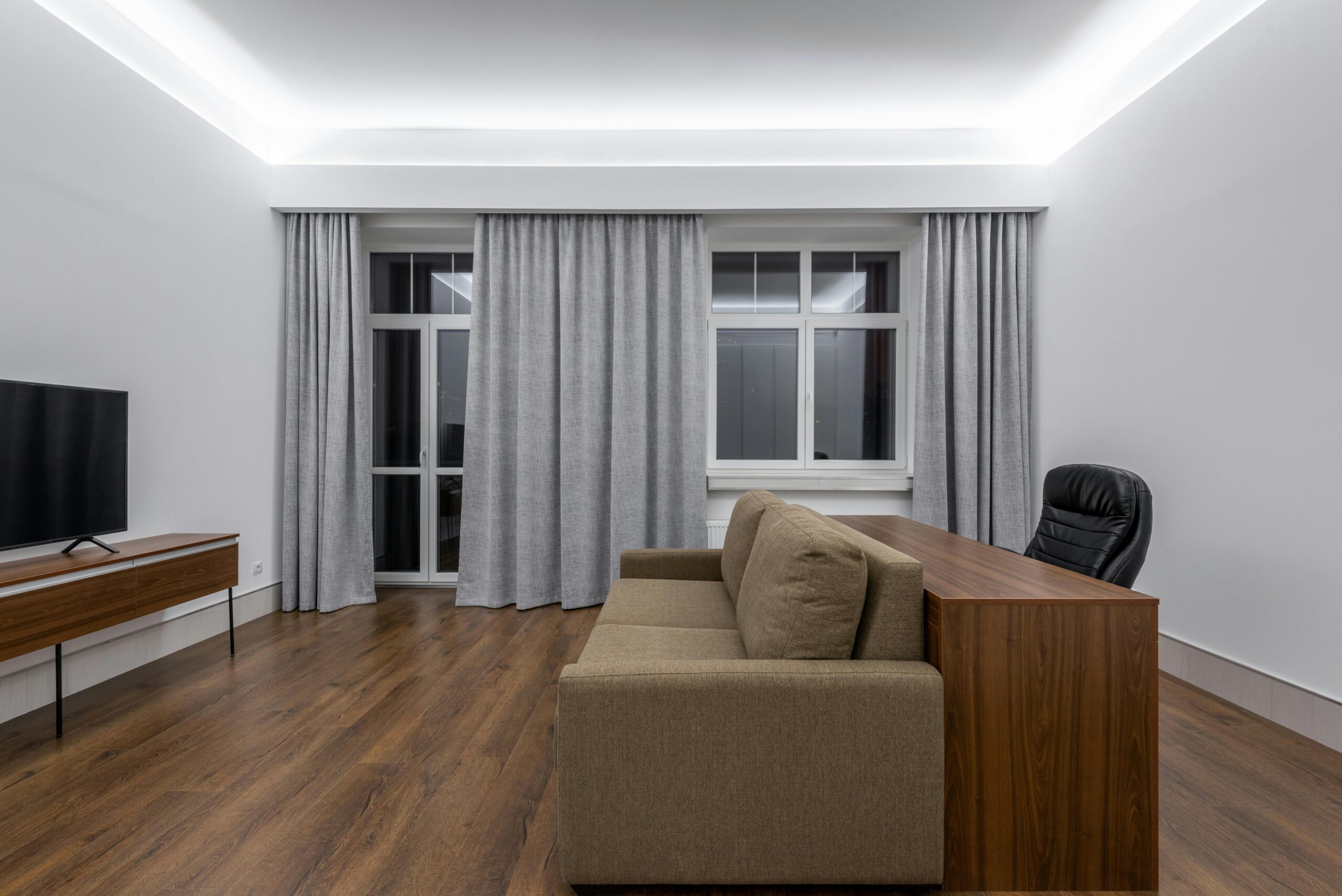 Wooden table near comfortable sofa against modern TV in spacious living room with curtains on window and minimalist design at home