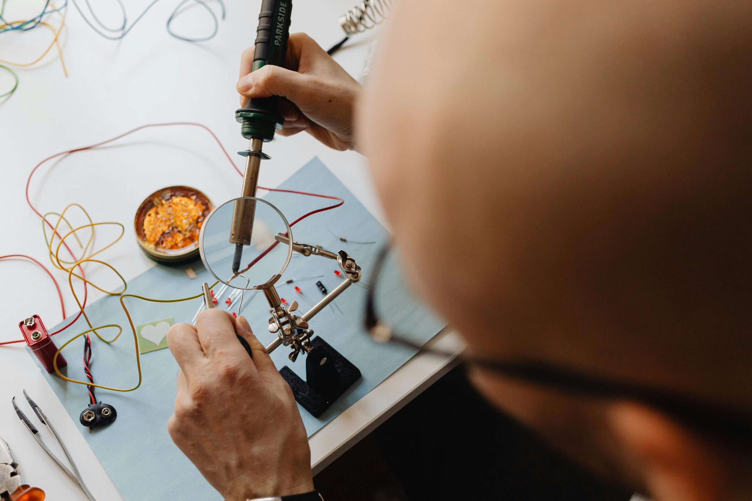Man soldering circuit board with a magnifying glass and soldering iron in a workshop setting.