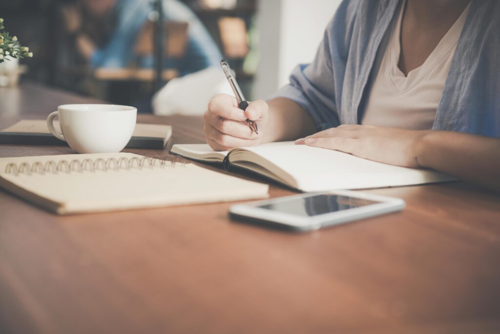 A woman writes in a notebook at a café table with a coffee and smartphone nearby. Zero-Based Budget