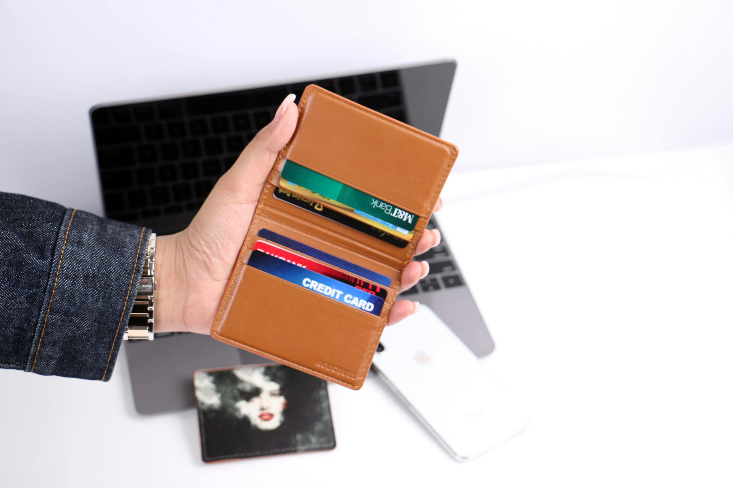 Close-up of a hand holding a leather wallet with credit cards, indoors with a laptop background.