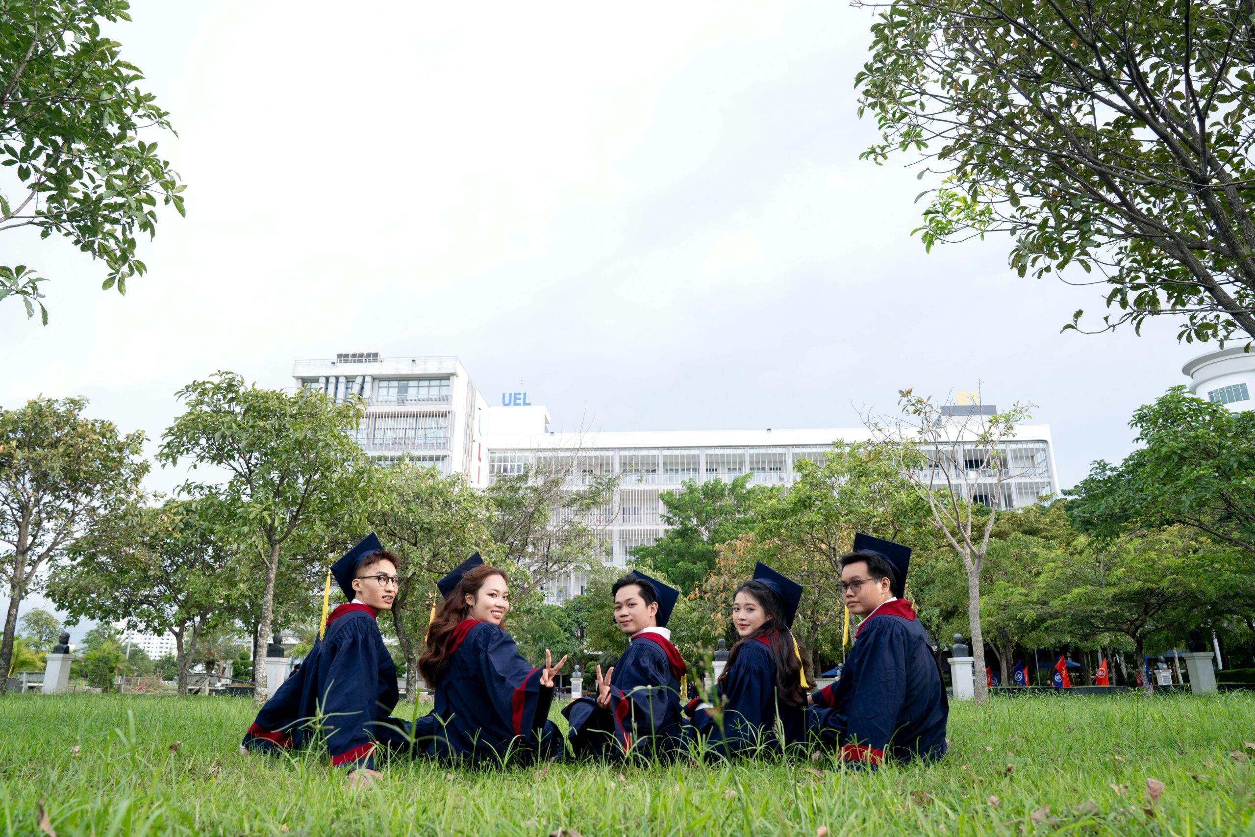 Group of five university graduates celebrating outdoors in regalia, sitting with a campus backdrop.