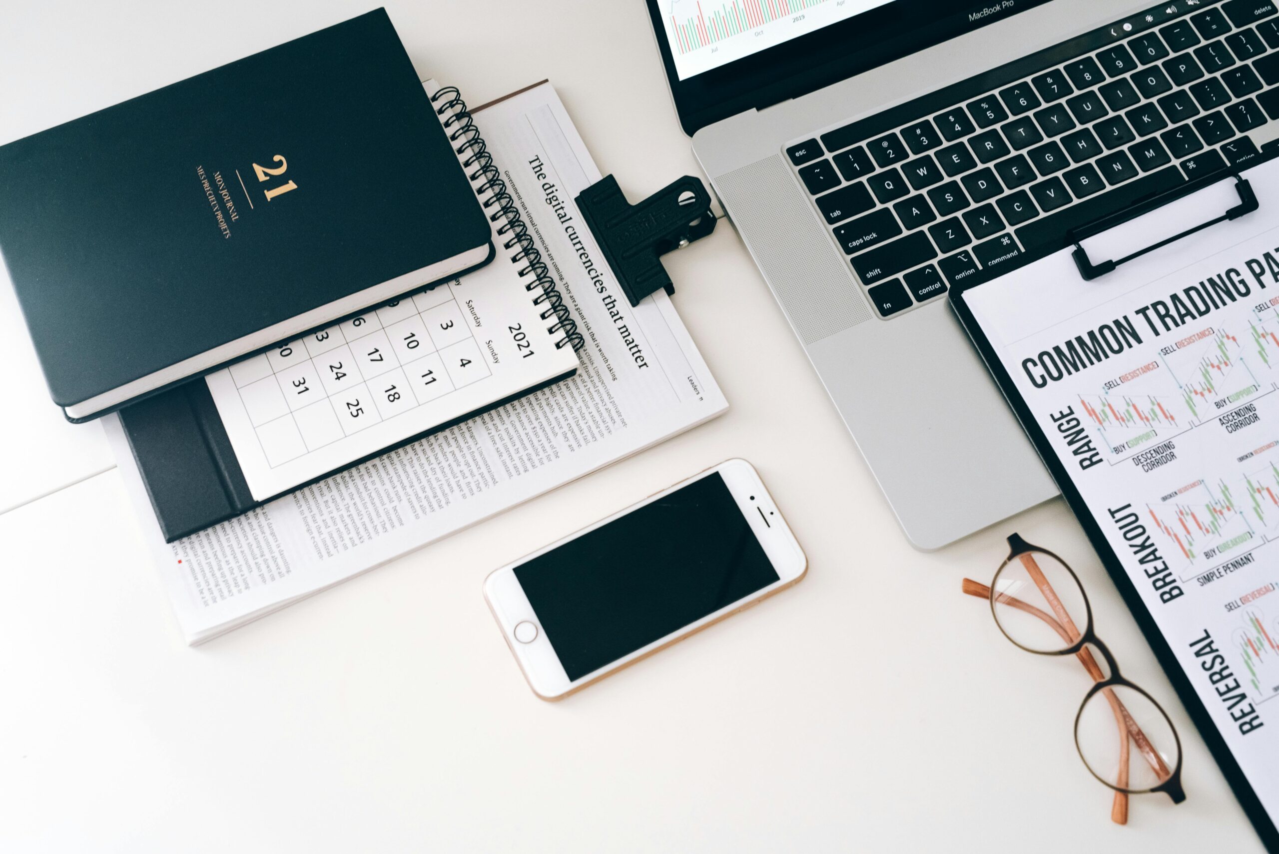 Flat lay of a modern workspace with a laptop, smartphone, planner, and trading charts on a white surface.