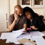 A couple sits at a table managing domestic finances, evaluating documents and using a smartphone.