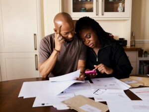 A couple sits at a table managing domestic finances, evaluating documents and using a smartphone.