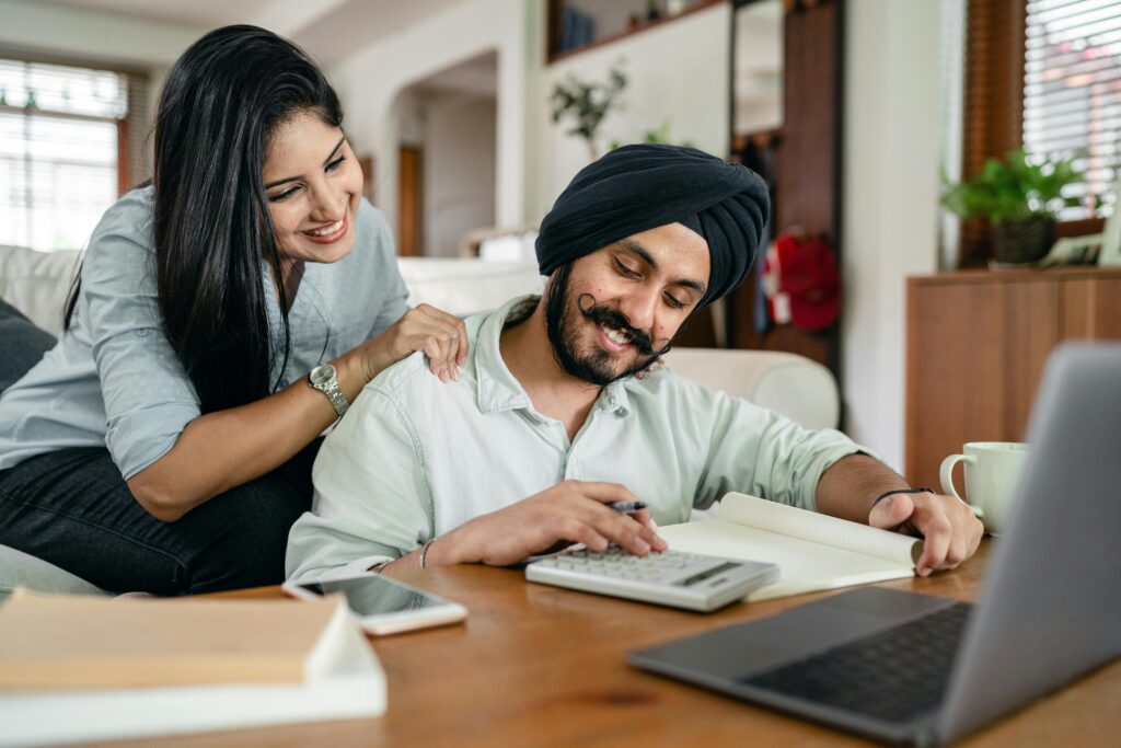 A smiling couple collaborates on financial paperwork at home, using a laptop and calculator. Manage Money as a Couple Without Fighting