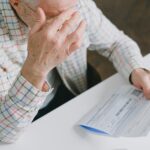 Elderly man in patterned shirt reading and holding bills at a home table, appearing focused.