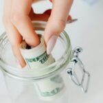 A close-up of a hand placing rolled dollars into a glass jar, symbolizing savings.