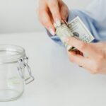 Woman rolling dollar bills beside a glass jar, symbolizing savings or financial planning.